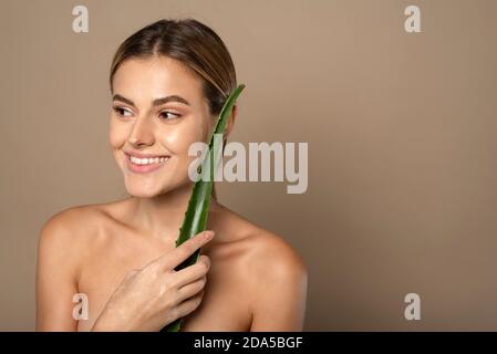 Sorridente giovane donna che tiene la foglia di aloe in mano su sfondo beige. Il concetto di cura della pelle, idratante con cosmetici naturali. Foto Stock
