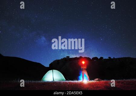 Uomo con faro rosso che guarda in telecamera vicino alla tenda di illuminazione, sullo sfondo cielo stellato sopra la montagna del Parco Nebrodi Foto Stock