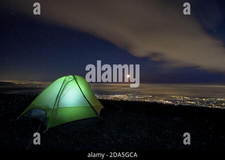 Una tenda illuminata ad alta quota nel Parco dell'Etna con vista sulla costa siciliana e sulla luna di notte cielo Foto Stock