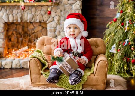 Un bambino piccolo in costume rosso di Babbo Natale, seduto sul divano della residenza, che tiene un regalo tra le mani. Carta anno nuovo. Il ragazzo biondo ride. Foto Stock