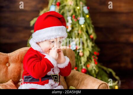 Un bambino piccolo in costume rosso di Babbo Natale, gioca nella sua residenza, lecca una palla di Capodanno come caramella. Cartolina festiva. Il ragazzo biondo ride. Natale Foto Stock