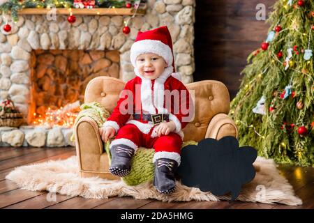 Un bambino piccolo in costume rosso di Babbo Natale, seduto sul divano della residenza, che tiene un regalo tra le mani. Carta anno nuovo. Il ragazzo biondo ride. Foto Stock