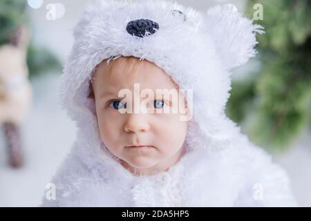 Un ragazzino vestito da orso polare bianco siede tra la foresta di alberi di Natale su una slitta di legno. Nelle vicinanze si trova una renna. Luci bokeh sfocate Foto Stock