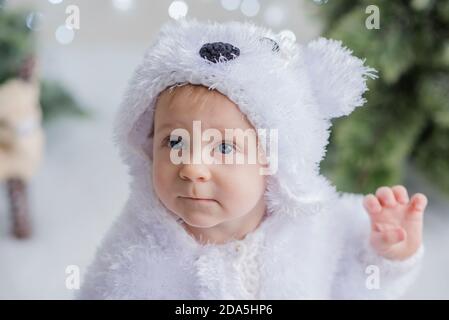 Un ragazzino vestito da orso polare bianco siede tra la foresta di alberi di Natale su una slitta di legno. Nelle vicinanze si trova una renna. Luci bokeh sfocate Foto Stock