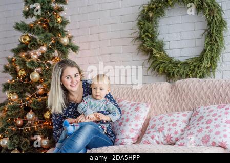 Una bella giovane donna si siede su un divano con un ragazzo vicino all'albero di Natale contro un muro di mattoni bianchi. Madre abbracci, baci, gioca con il suo piccolo figlio. Felice Foto Stock