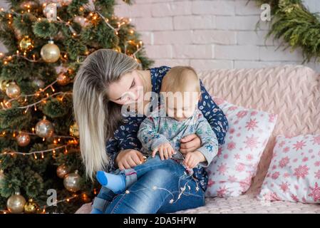 Una bella giovane donna si siede su un divano con un ragazzo vicino all'albero di Natale contro un muro di mattoni bianchi. Madre abbracci, baci, gioca con il suo piccolo figlio. Felice Foto Stock