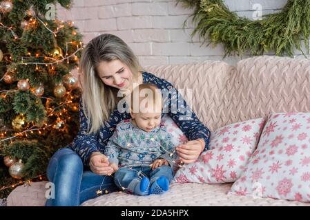 Una bella giovane donna si siede su un divano con un ragazzo vicino all'albero di Natale contro un muro di mattoni bianchi. Madre abbracci, baci, gioca con il suo piccolo figlio. Felice Foto Stock