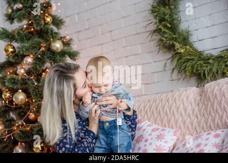 Una bella giovane donna si siede su un divano con un ragazzo vicino all'albero di Natale contro un muro di mattoni bianchi. Madre abbracci, baci, gioca con il suo piccolo figlio. Felice Foto Stock