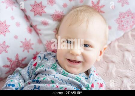 Un bambino piccolo in costume rosso di Babbo Natale, seduto sul divano della residenza, che tiene un regalo tra le mani. Carta anno nuovo. Il ragazzo biondo ride. Foto Stock