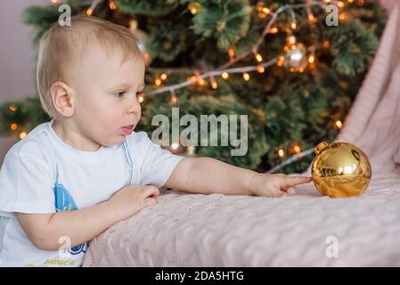 Piccolo biondo ragazzo gioca con una palla d'oro all'albero di Natale a casa. Tessera Capodanno. Primo piano ritratto del Kid ridendo, toccando il giocattolo Foto Stock