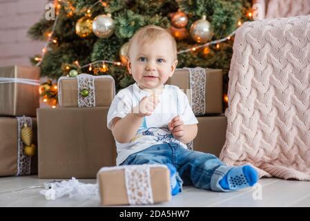 Ragazzo biondo che mangia un uomo di pan di zenzero vicino all'albero di Natale a casa. Carta per vacanze di Capodanno con paludi bokeh. Il bambino ride, apre una confezione regalo Foto Stock