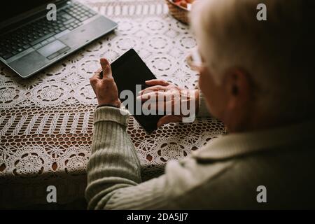 Primo piano di una donna caucasica anziana mano mentre digitando su un tablet, mentre di fronte a lei sul tavolo è un computer portatile durante la quarantena COVID - 19 Foto Stock