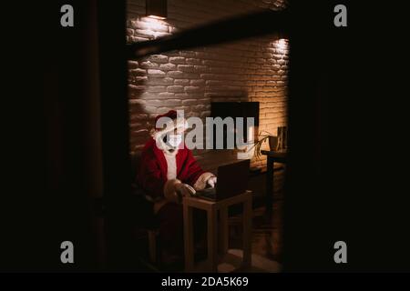 Babbo Natale con una maschera davanti a un computer portatile e che parla in una videochiamata in camera. Celebrazione del nuovo anno e Natale Foto Stock
