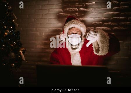 Un ritratto di un uomo caucasico vestito in costume di Babbo Natale seduto di fronte a un computer portatile e dando un pollice giù. Festa di Capodanno Foto Stock
