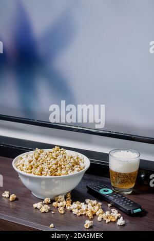 Una ciotola piena di popcorn, alcuni popcorn caduti sul tavolo accanto ad una lager davanti ad una tv LED con un telecomando. Foto Stock