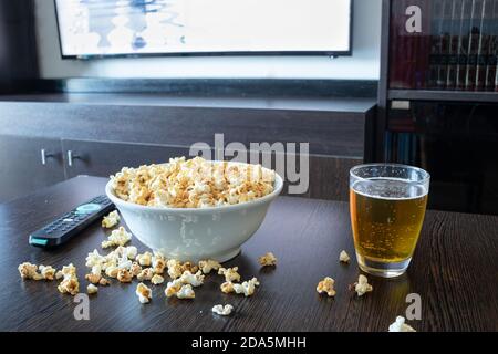 Una ciotola piena di popcorn, alcuni popcorn caduti sul tavolino accanto ad una lager davanti ad una tv LED con telecomando. Foto Stock