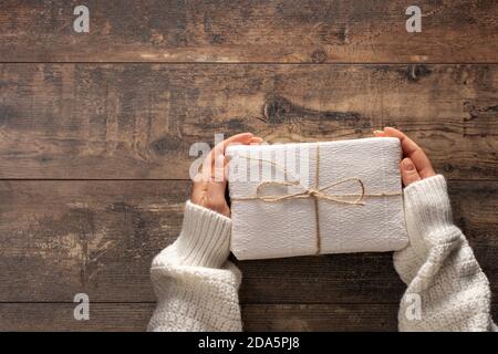 donna che tiene la scatola del regalo di natale. Tavolo rustico in legno. Vista dall'alto mani donna che tengono presente la scatola. Mano femmina che tiene scatola regalo su sfondo tavolo di legno Foto Stock