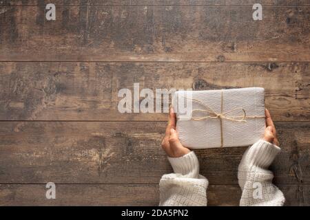 Vista dall'alto sulle mani di una giovane donna che tiene Natale, compleanno o qualsiasi altro regalo di celebrazione avvolto in carta bianca su rustico sfondo di legno. Preparazione Foto Stock