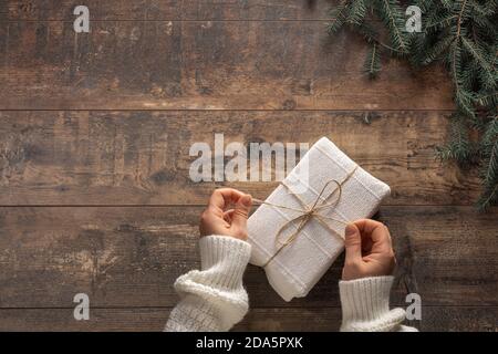 Vista dall'alto sulle mani di una giovane donna che tiene in mano la confezione regalo di Natale avvolta in carta bianca su rustico sfondo di legno. Preparazione per le vacanze e la celebrazione. Foto Stock