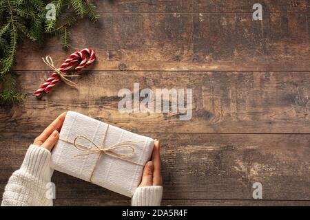 Confezione regalo regalo Natale in mani di donna su sfondo rustico in legno. Rami dell'albero di Natale e canne caramelle su vecchio tavolo in legno. Copia spac Foto Stock