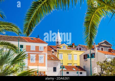 Bellissima città di Lussino sull'isola di Lussino, la costa adriatica in Croazia, la torre della cattedrale e il centro della città, vista attraverso le foglie di palma Foto Stock