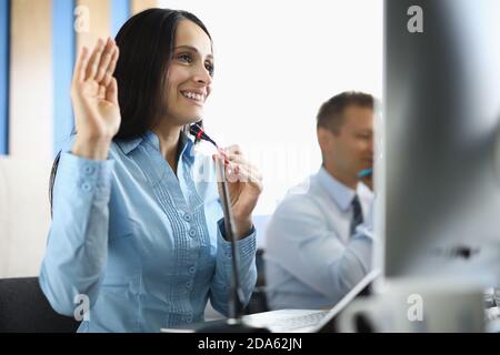 Donna d'affari si siede al computer con il microfono sorridente e sollevando la mano in modo accogliente. Foto Stock