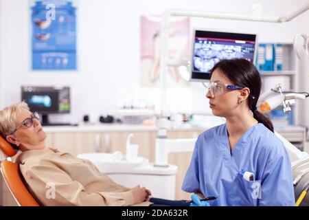 Assistente nel gabinetto dentista durante la donna anziana igiene orale controllare seduta su sedia. Donna anziana che parla con l'infermiera medica in ufficio di stomatologia circa il problema dei denti. Foto Stock