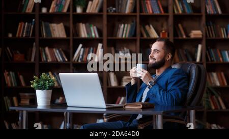 Caffè fresco al lavoro. Giovane sicuro in abbigliamento casual elegante che lavora su un computer portatile e beve una bevanda calda mentre si siede al lavoro Foto Stock