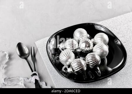 Impostazione del tavolo di Natale. Palle di albero di Natale su un piatto nero, coltello, forchetta. Stoviglie nere. Il concetto di Natale e il nuovo anno. Composizione monocromatica in argento minimalista Foto Stock
