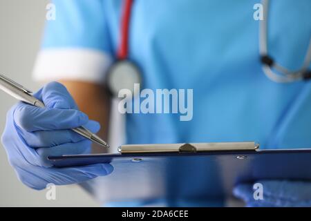 La mano del medico nei guanti tiene la penna e compila i documenti sulla clipboard. Foto Stock