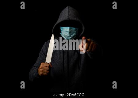 Il giovane uomo in maschera medica punta il dito mentre tiene il coltello su sfondo nero Foto Stock