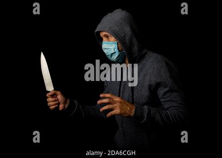 Un giovane aggressivo in una maschera medica in un cappuccio con un coltello su sfondo nero Foto Stock