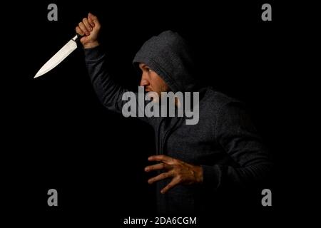 Un giovane aggressivo in una cappa branchezza un coltello contro uno sfondo nero Foto Stock