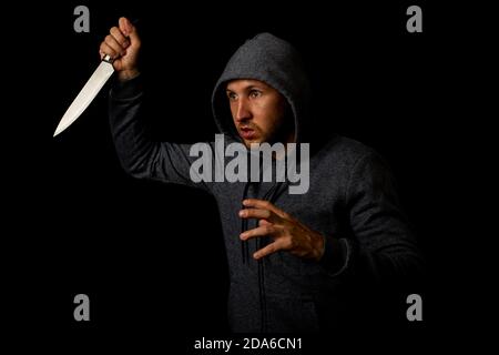 Un giovane aggressivo in una felpa in un cappuccio con un coltello su sfondo nero Foto Stock