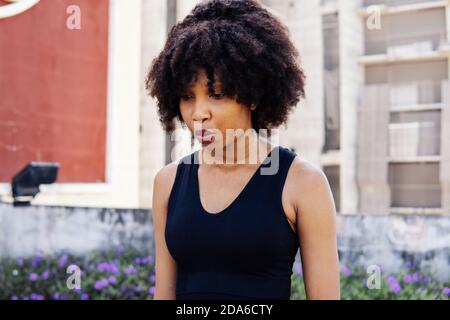 Giovane donna sportiva afroamericana che si riposa dopo l'allenamento fitness. Fitness atleta femminile esausta e stanca guardando lontano e in piedi nel parco urbano della città Foto Stock