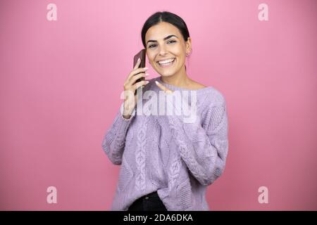 Bella donna che indossa un maglione viola casual su sfondo rosa sorridente, indicando il telefono che ha nell'orecchio Foto Stock