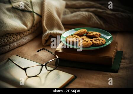 Sul letto si trovano vecchi libri, maglioni caldi, bicchieri neri e un piatto di dolci biscotti al cioccolato. Comfort a casa. Tempo di lettura. Tempo libero. Foto Stock