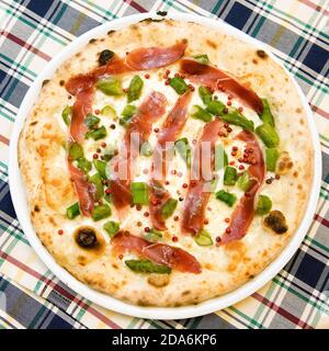 una vera e propria pizza italiana originale in un piatto, pepe rosa, prosciutto, asparagi, mozzarella e basilico, vista dall'alto su una plaid tovaglia Foto Stock