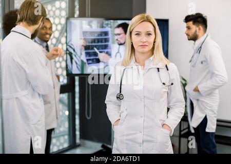 Attraente donna bionda matura medico, in posa a macchina fotografica. Colleghi multirazziali vicino grande schermo su sfondo, discutendo la TAC del paziente e. Foto Stock