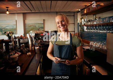 Ritratto di una bella giovane proprietaria di caffè in piedi dietro il bancone indossare un grembiule che sorregge il tablet digitale Foto Stock