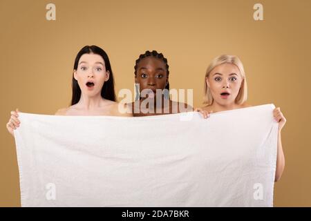 Tre donne multietniche emotive nascondono dietro asciugamano da bagno con scioccato Espressione del volto Foto Stock