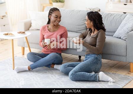 Le amiche nere felici si divertono il fine settimana insieme, bevendo il caffè sul divano Foto Stock