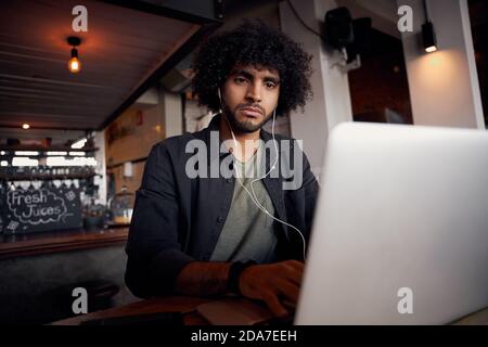 Triste giovane maschio guardare video su computer portatile con auricolari in caffetteria moderna Foto Stock