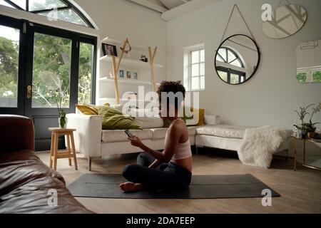 Giovane donna africana che usa il cellulare mentre si siede sul tappeto di yoga a casa Foto Stock