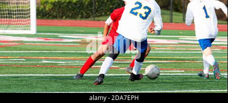 Vista posteriore di un giocatore di calcio in una maglia bianca che dribbling la palla intorno al suo avversario in una maglia rossa durante una partita di scuola superiore su un campo di erba verde. Foto Stock