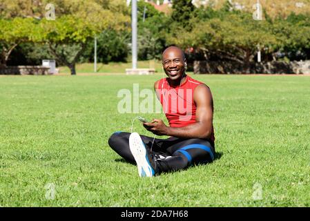 Ritratto di giovane uomo sportivo africano rilassato seduto all'aperto prato e ascoltare musica Foto Stock
