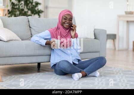 Giovane donna musulmana nera godendo del tè e parlando al telefono A casa Foto Stock