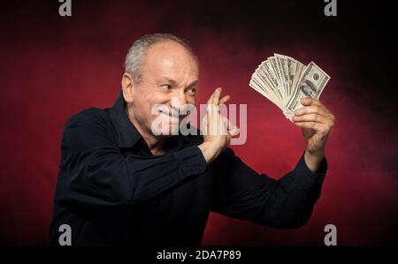 Signore anziano che tiene una pila di soldi. Ritratto di un vecchio eccitato. Felice vecchio uomo che tiene le banconote del dollaro su sfondo rosso scuro. Foto Stock