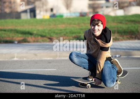 Skateboarder maschile seduto sullo skateboard mentre si guarda la fotocamera all'aperto Foto Stock