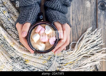 Coppa per mani femminili con bevanda calda con marshmallows su tavolo di legno con sciarpa di lana a maglia calda. Natale sfondo - il concetto di inverno, calore, vacanze ed eventi.Soft focus.Vista dall'alto Foto Stock